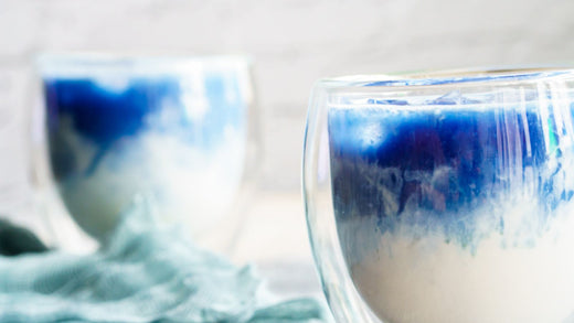 Two double-walled glass cups filled with Blue Serenity Latte, featuring a gradient of blue and white swirls. A gold spoon with vibrant BlueMAJIK powder sits in the foreground, accompanied by a soft teal cloth draped in the background.