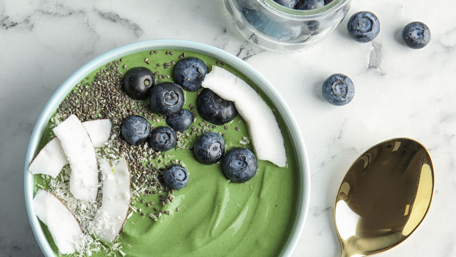 A green smoothie bowl topped with fresh blueberries, coconut flakes, and chia seeds. A jar of blueberries and scattered loose berries are visible in the background, accompanied by a shiny gold spoon on the side.