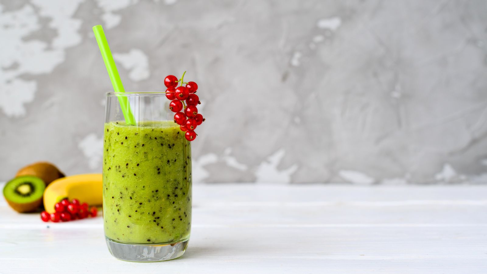 A vibrant green E3AFA smoothie served in a clear glass garnished with a cluster of red currants. In the background, a whole kiwi, banana, and additional red currants.