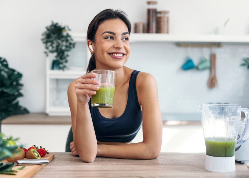 A smiling woman in a modern kitchen holding a glass of green smoothie, with a blender and fresh fruits visible on the counter, symbolizing health and wellness.