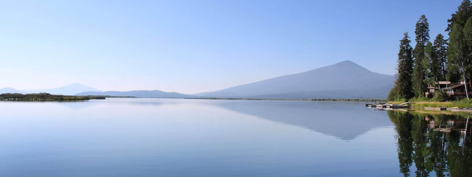 A serene view of Klamath Lake in Oregon, featuring calm waters that reflect the clear blue sky and surrounding landscape. In the background, a mountain range is visible, adding depth to the scene. On the right side, a cluster of tall evergreen trees lines the lake’s shore, with wooden docks extending into the water and a few lakeside houses nestled among the trees. The atmosphere is peaceful, with no visible human activity, showcasing the lake’s natural beauty.