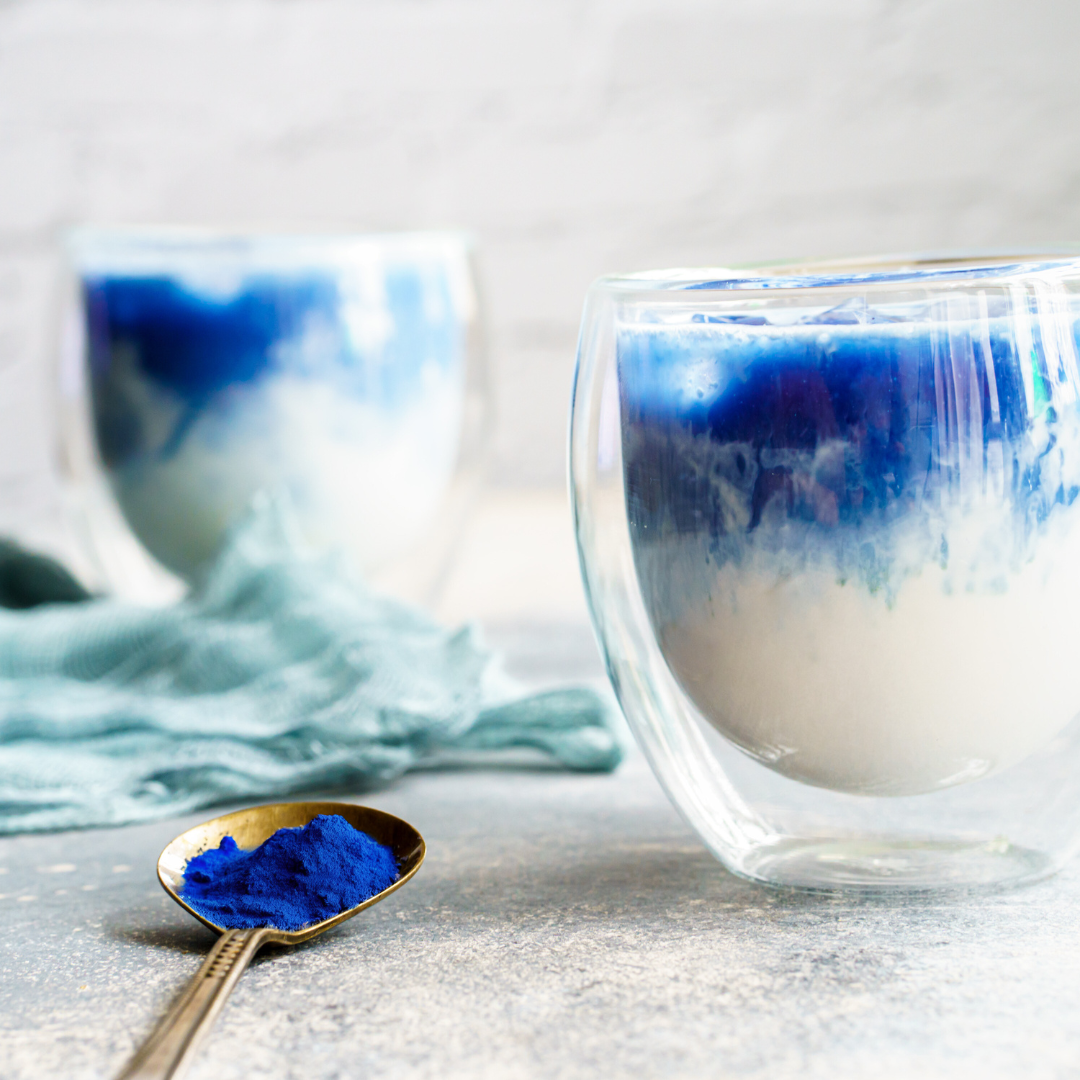 Two glasses of a vibrant blue BlueMAJIK drink, showcasing the antioxidant-rich superfood in a creamy blend, with a spoon of BlueMAJIK powder in the foreground.