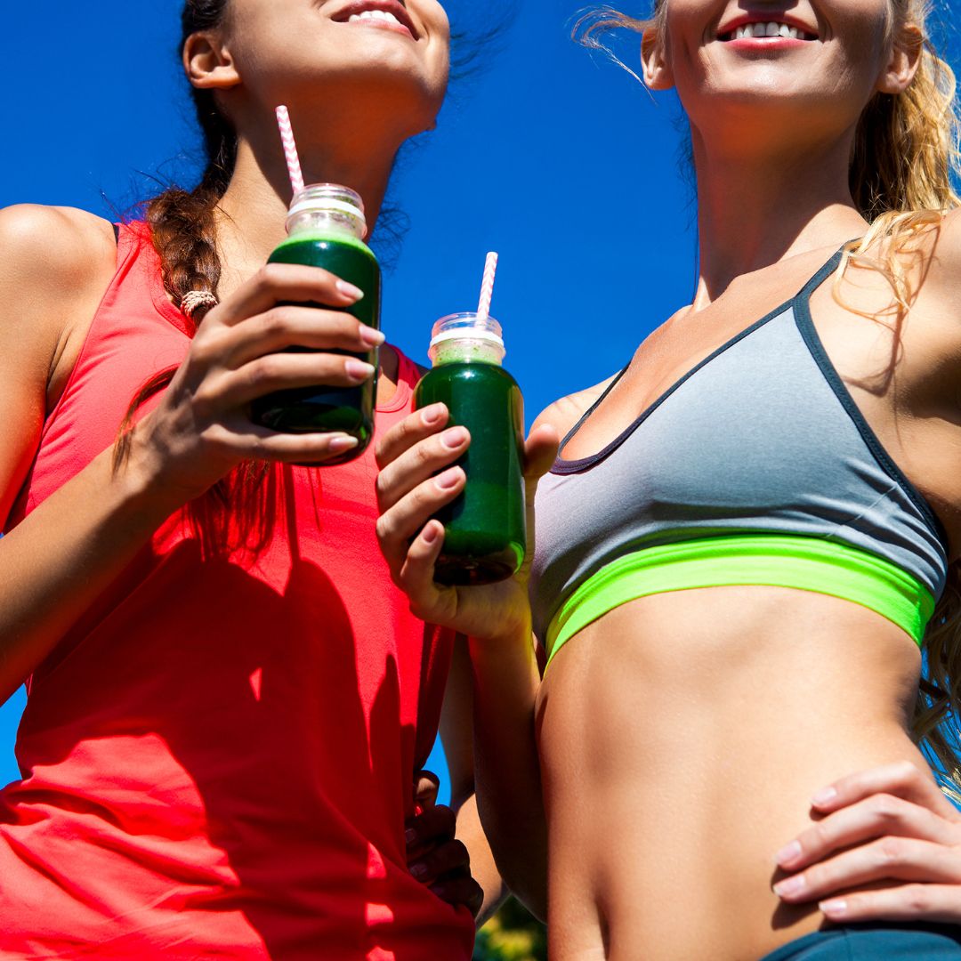 Two fit and active women wearing athletic wear, holding glass bottles filled with a vibrant green smoothie, likely infused with E3AFA (Aphanizomenon flos-aquae). They are smiling and standing under a bright blue sky, symbolising health, vitality, and wellness. The image conveys energy, hydration, and the benefits of superfoods for an active lifestyle.