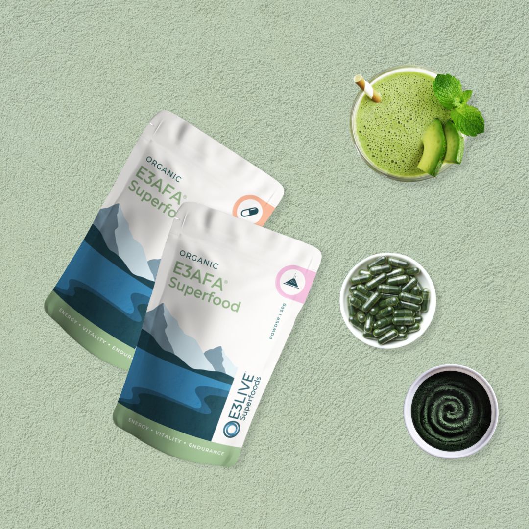 Two packs of E3AFA® Superfood, one labeled for capsules and the other for powder. Surrounding the packs are a green smoothie garnished with avocado and mint, a bowl of green capsules, and a swirl of green powder representing the nutrient-rich E3AFA® and E3Live® products.