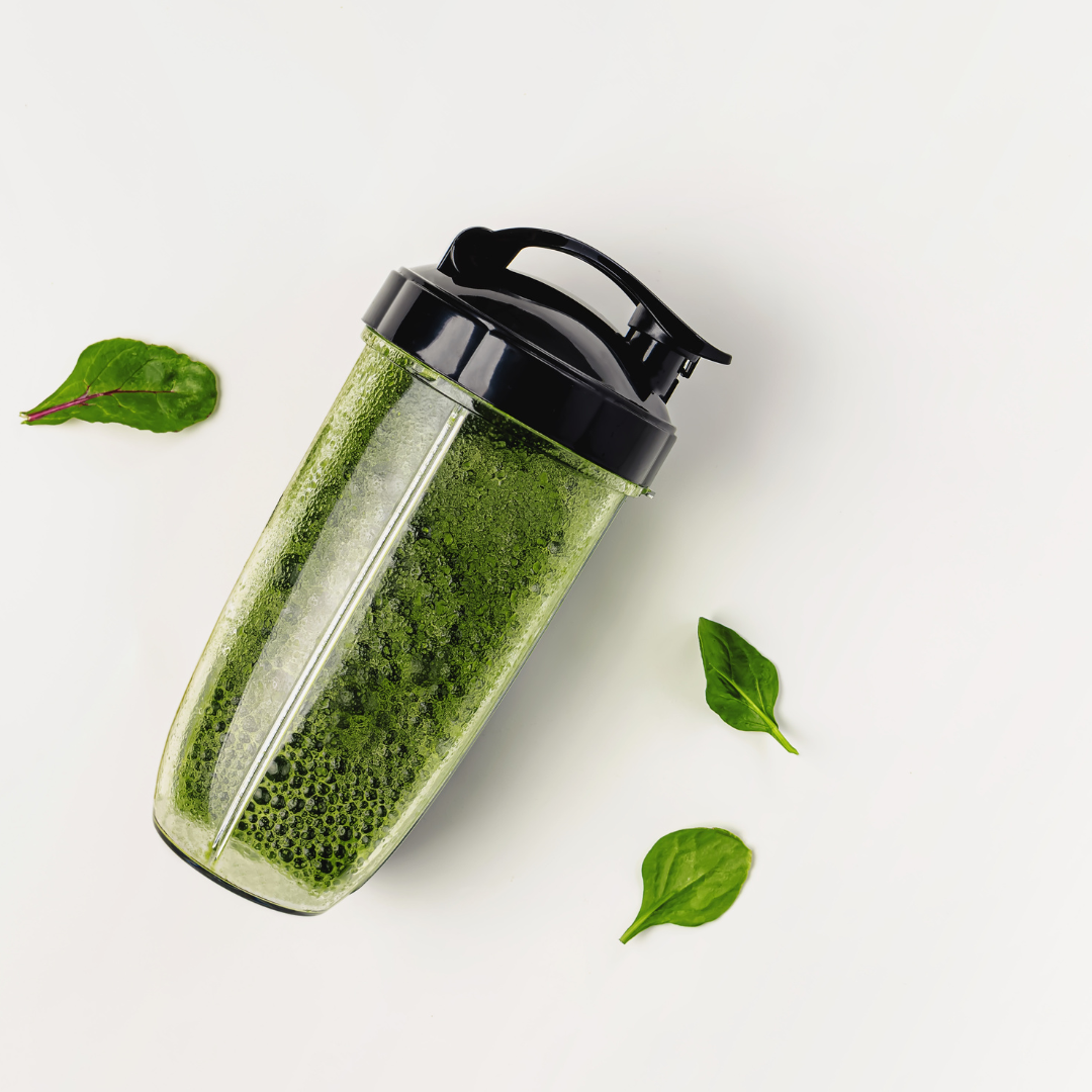 A green smoothie made with E3AFA powder in a transparent shaker bottle with a black lid, lying on a white surface. The smoothie has a rich green color with visible froth and bubbles. Fresh spinach leaves are scattered around the bottle, enhancing the natural and healthy appeal of the drink. The clean, minimalistic background highlights the vibrant colour and freshness of the smoothie.