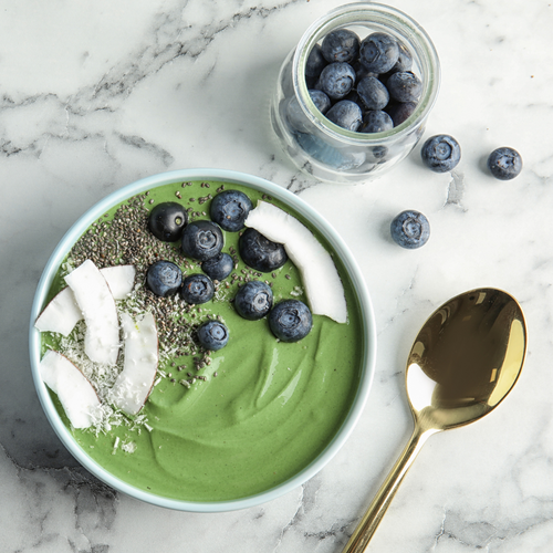 A green smoothie bowl topped with fresh blueberries, coconut flakes, and chia seeds. A jar of blueberries and scattered loose berries are visible in the background, accompanied by a shiny gold spoon on the side.