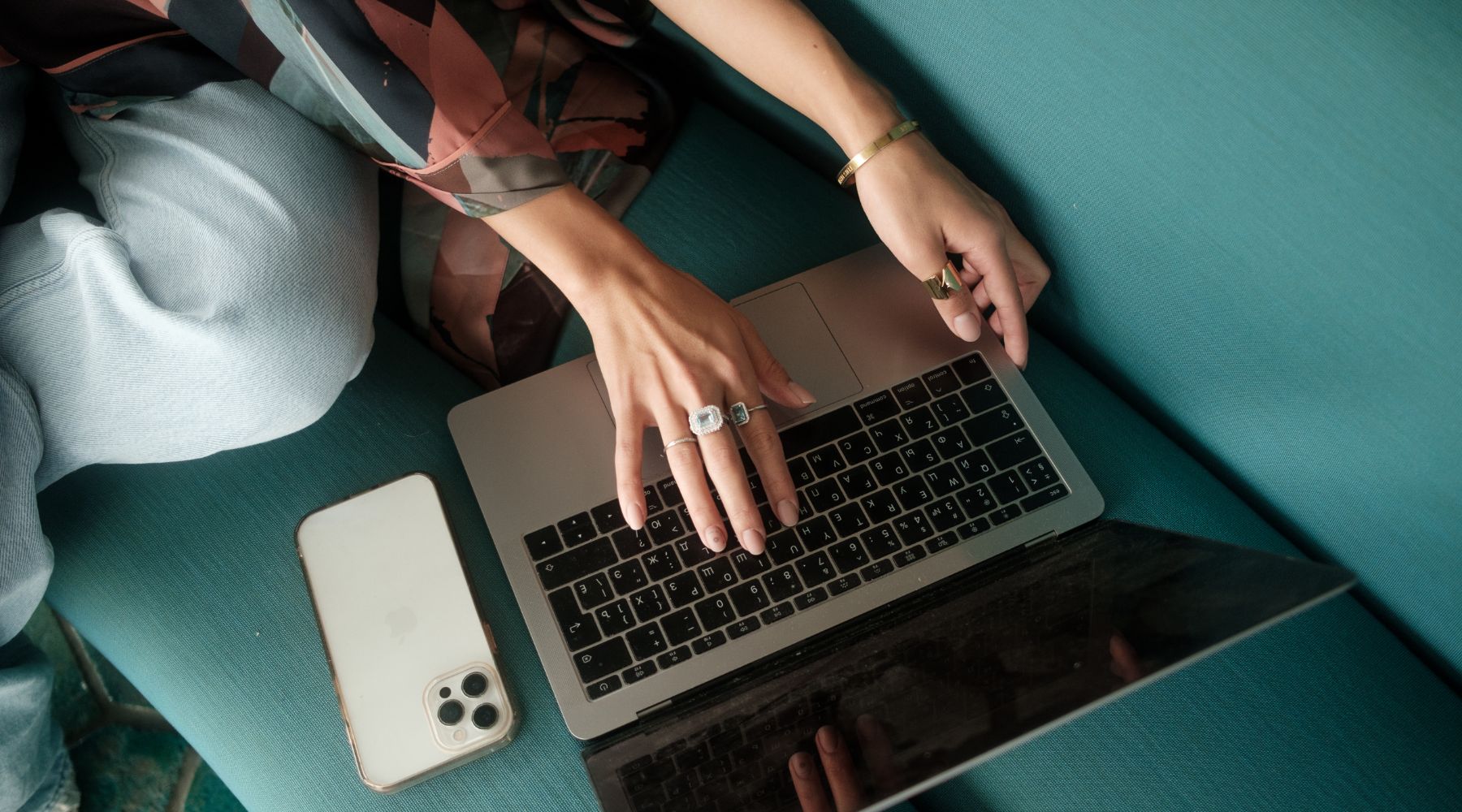 Close-up of a person using a laptop on a teal sofa, with hands adorned in rings and bracelets. An iPhone lies beside them, enhancing a focused, modern workspace aesthetic. This image reflects a lifestyle that may benefit from Organic BrainON Superfood, suggesting mental clarity, productivity, and a balanced, health-conscious approach to daily tasks.